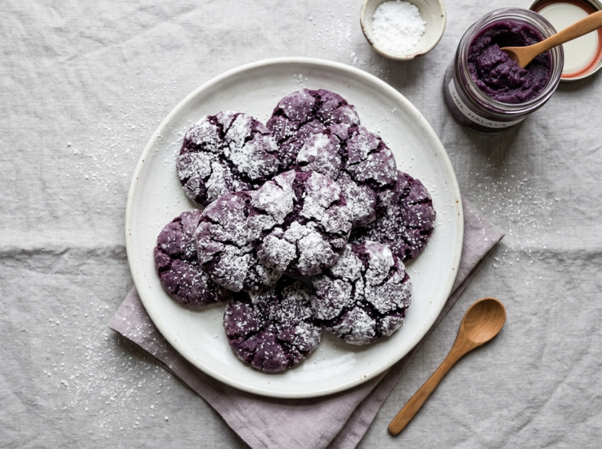 Batch of ube crinkle cookies with deep purple tops and powdered sugar cracks on a white plate, jar of ube halaya nearby