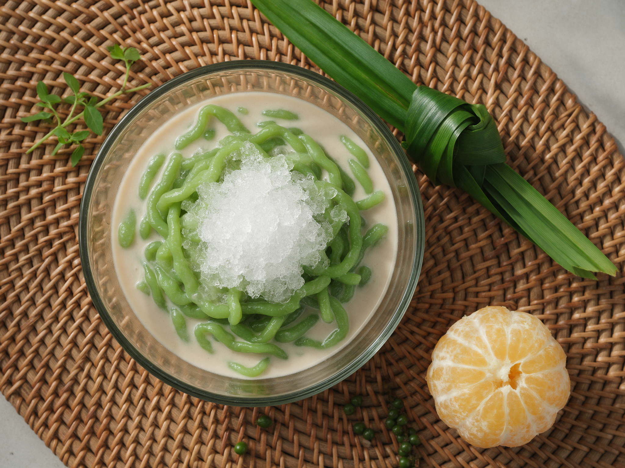 Overhead view of green pandan lod chong noodles in coconut milk with crushed ice in a glass bowl on a rattan mat